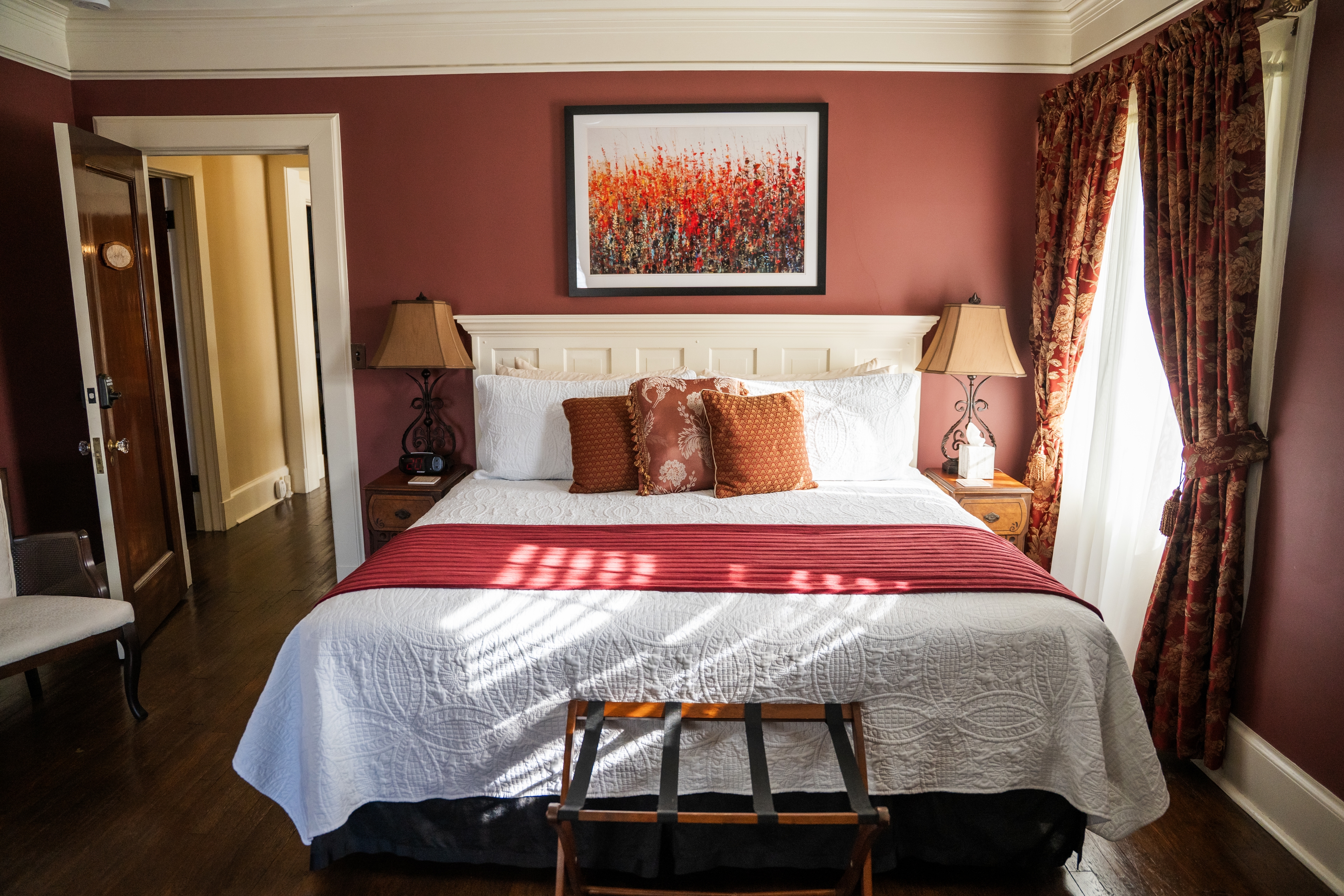 A sunlit bedroom with deep red walls and dark hardwood floors, featuring a white bed with a red blanket abstract orange wall art, and patterned curtains.