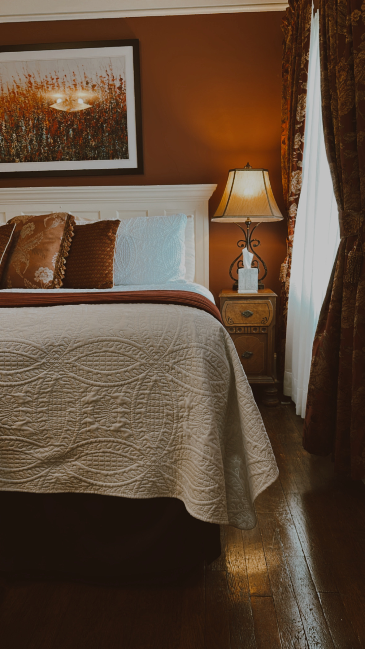 A close-up of a cozy bed with a white quilted bedspread and a red accent runner, featuring burnt-orange pillows and a matching abstract painting above the headboard.
