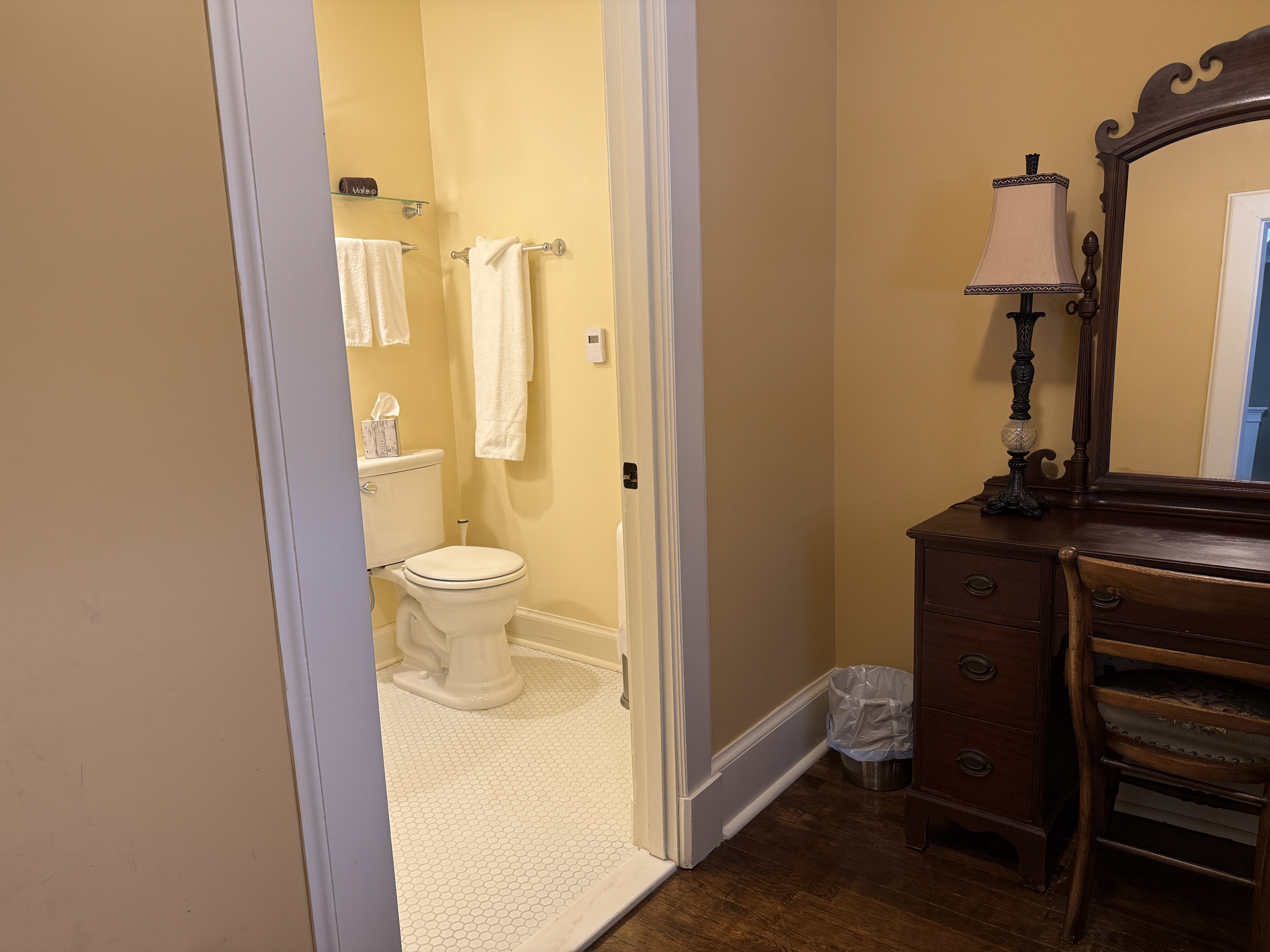 A view from a bedroom into a bright, neutral-toned bathroom featuring a white toilet, white tiled floors, and fresh white towels hanging on a silver rack.