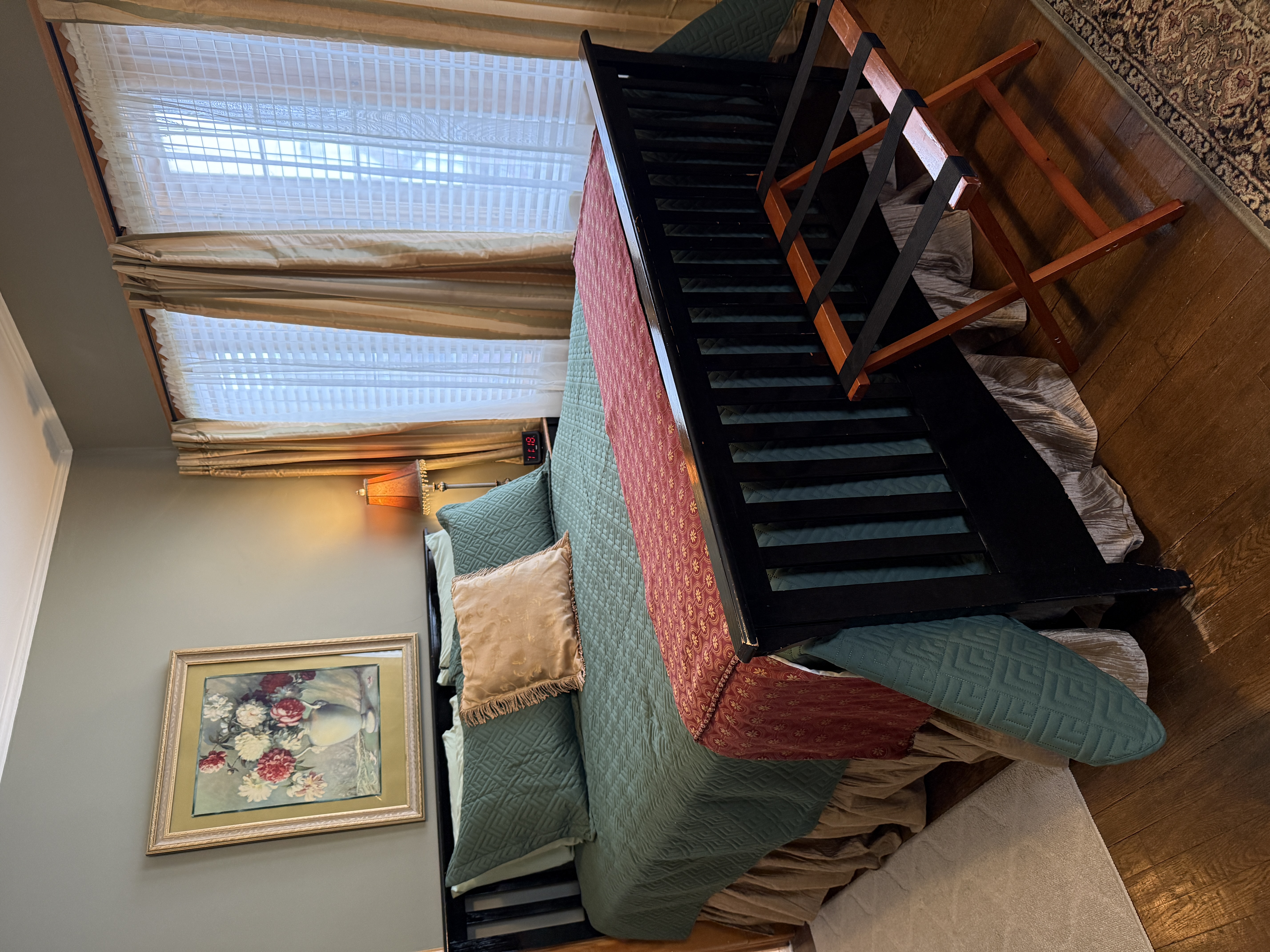 A bedroom with grey-green walls and dark hardwood floors featuring a black slatted bed frame, a teal quilted bedspread with a red patterned throw, and framed floral artwork above the headboard.