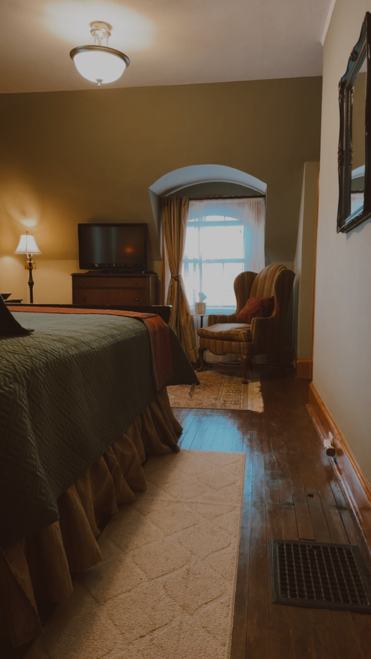 A cozy bedroom area with grey-green walls and dark hardwood floors, featuring a teal quilted bed and a tan wingback chair tucked into a sunlit arched window nook.
