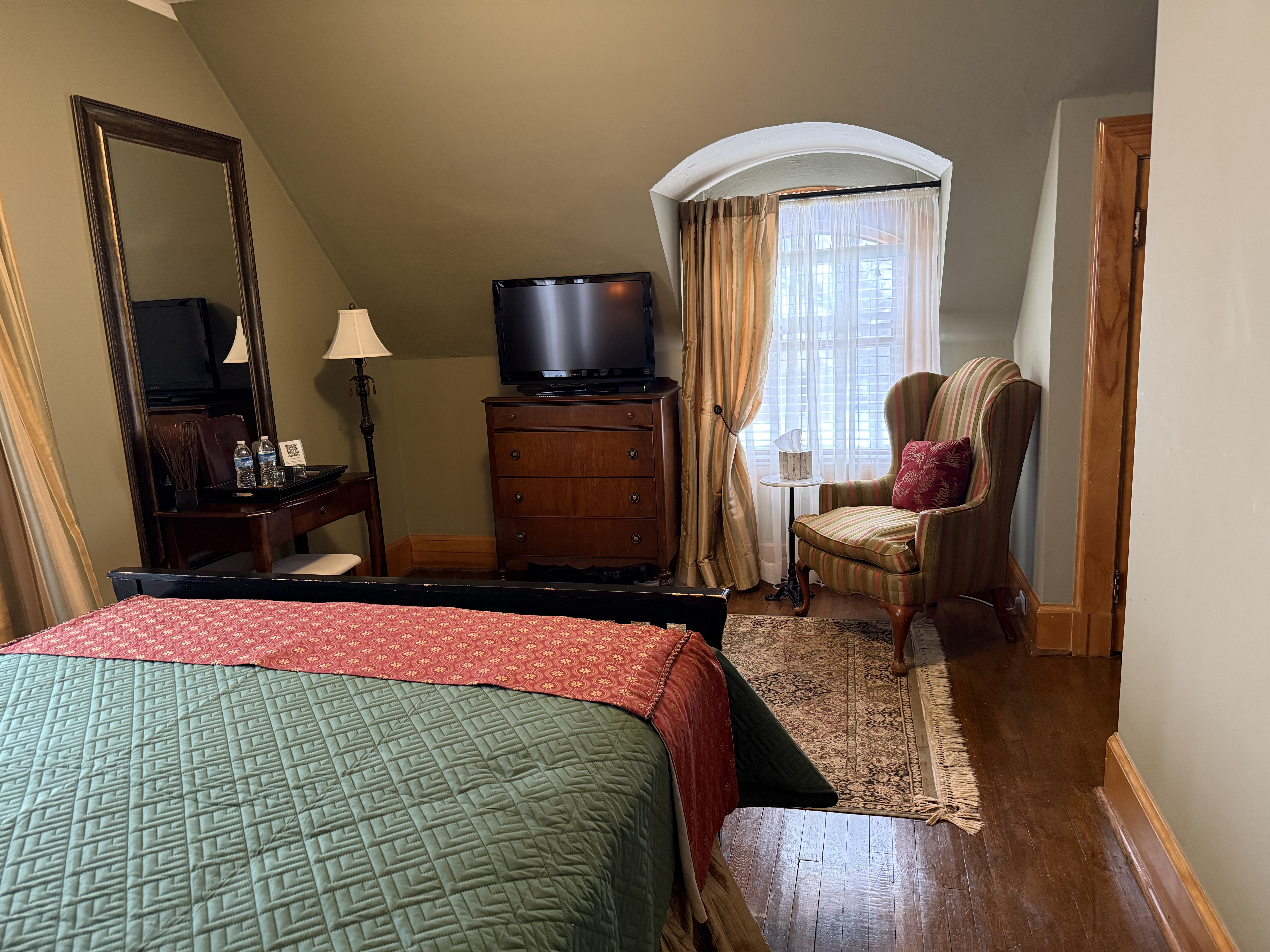 A cozy bedroom nook with olive-green walls and a sloped ceiling, featuring a flat-screen TV on a dark wood dresser, a tan wingback chair by an arched window, and a glimpse of a teal quilted bed.