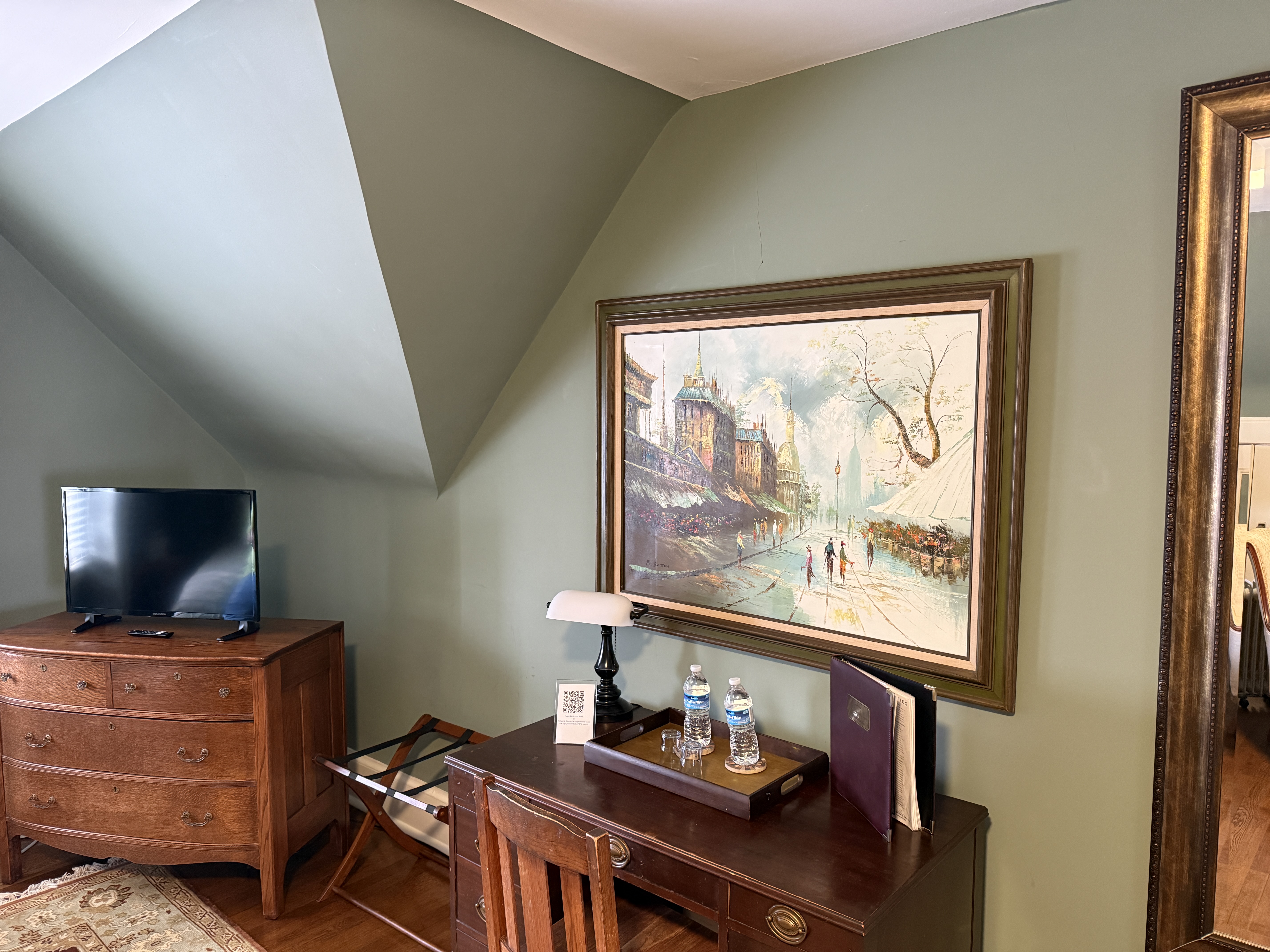 A cozy work area within a room featuring sage green walls and sloped ceilings, showcasing a dark wood desk with a vintage-style lamp, a large gold-framed painting of a European street scene, and a classic wood dresser with a flat-screen TV.