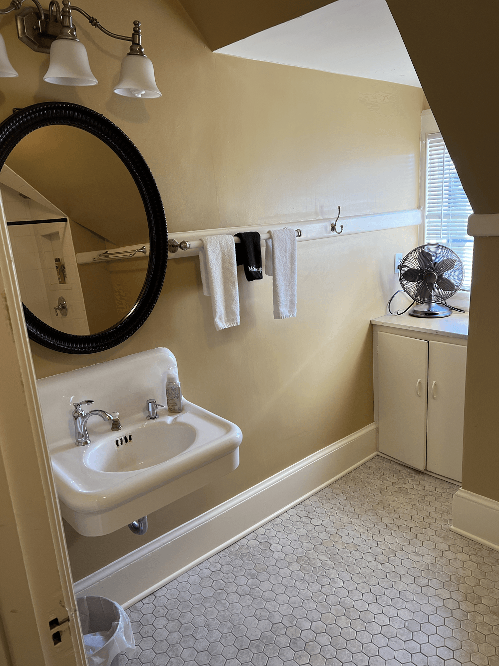 A classic bathroom under a sloped ceiling with tan walls and grey penny tile heated floor. It features a white wall-mounted sink, a round black-framed mirror, and a small white built-in cabinet tucked into the corner by a window.