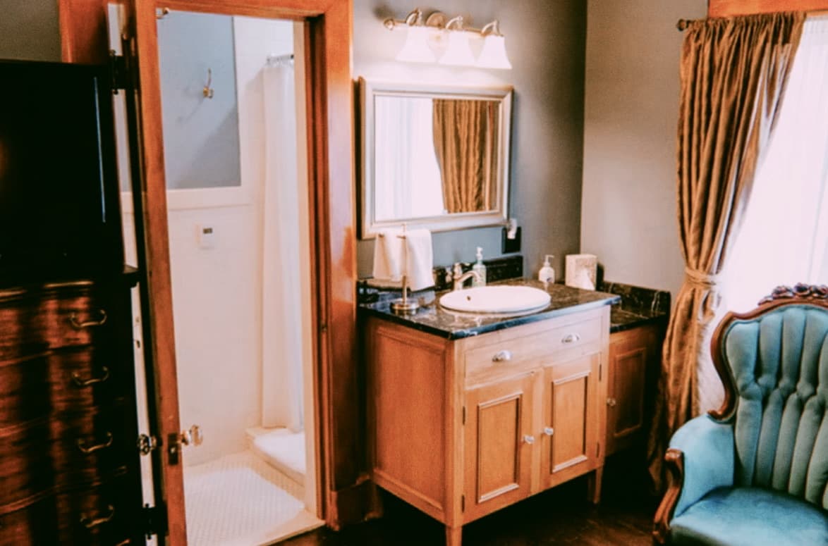 A vanity area featuring a light wood cabinet with a black stone countertop, a silver-framed mirror with overhead lighting, and a teal velvet armchair beside a window.