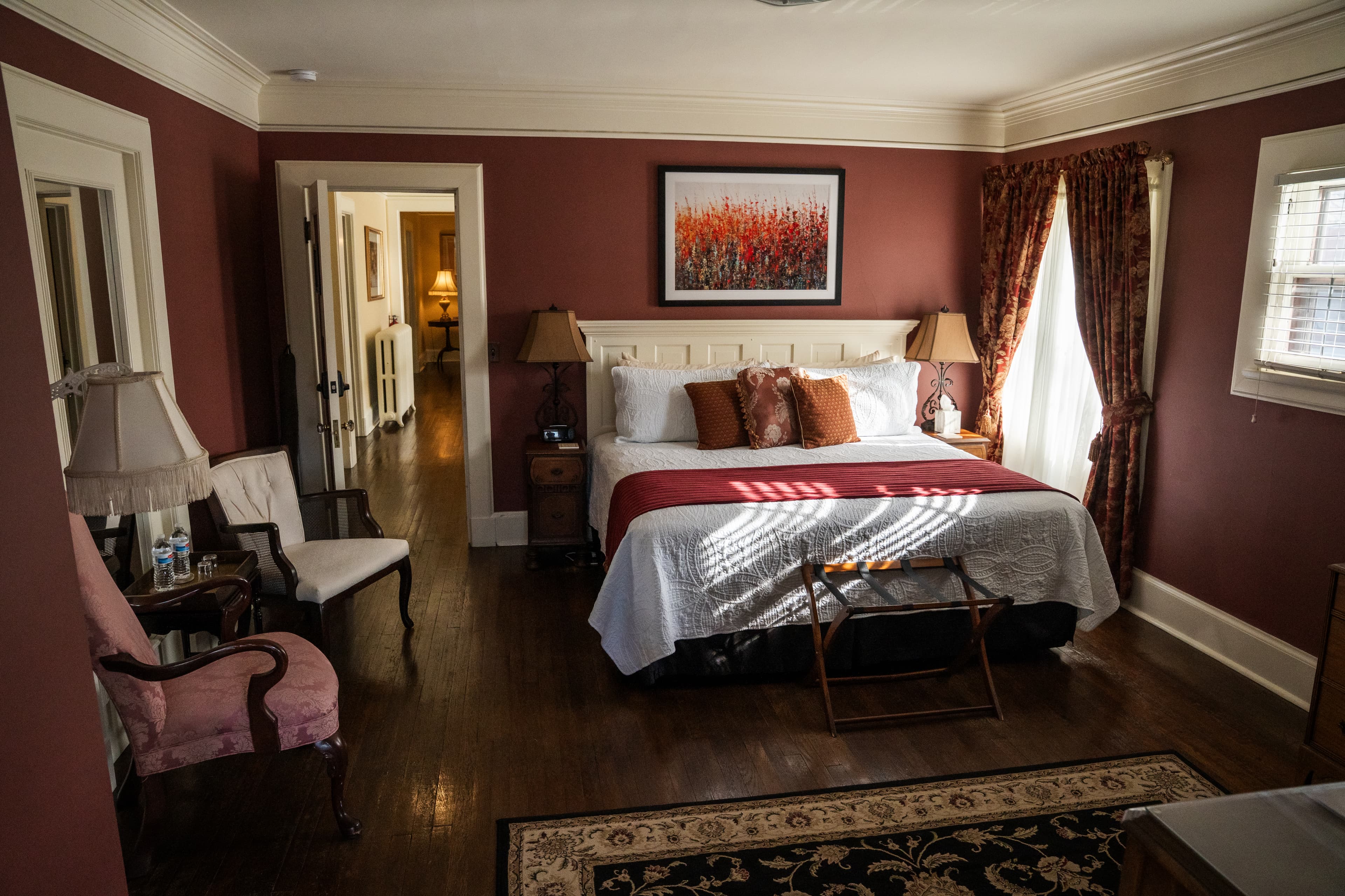 An elegant bedroom with deep red walls, featuring a white headboard bed with a red accent throw, a vintage pink armchair, hardwood floors, and a decorative area rug.