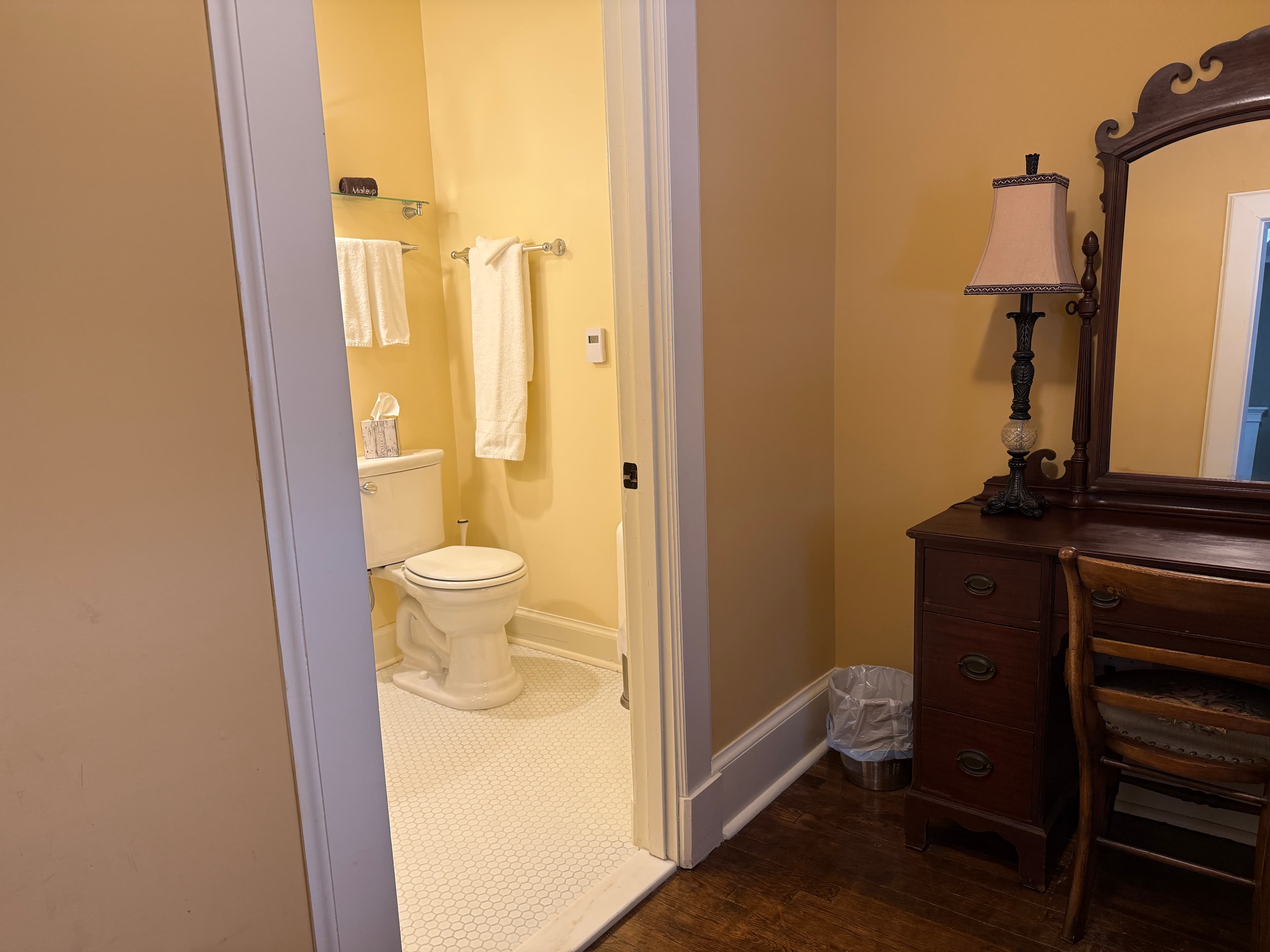 A view from a bedroom into a bright, neutral-toned bathroom featuring a white toilet, white tiled floors, and fresh white towels hanging on a silver rack.
