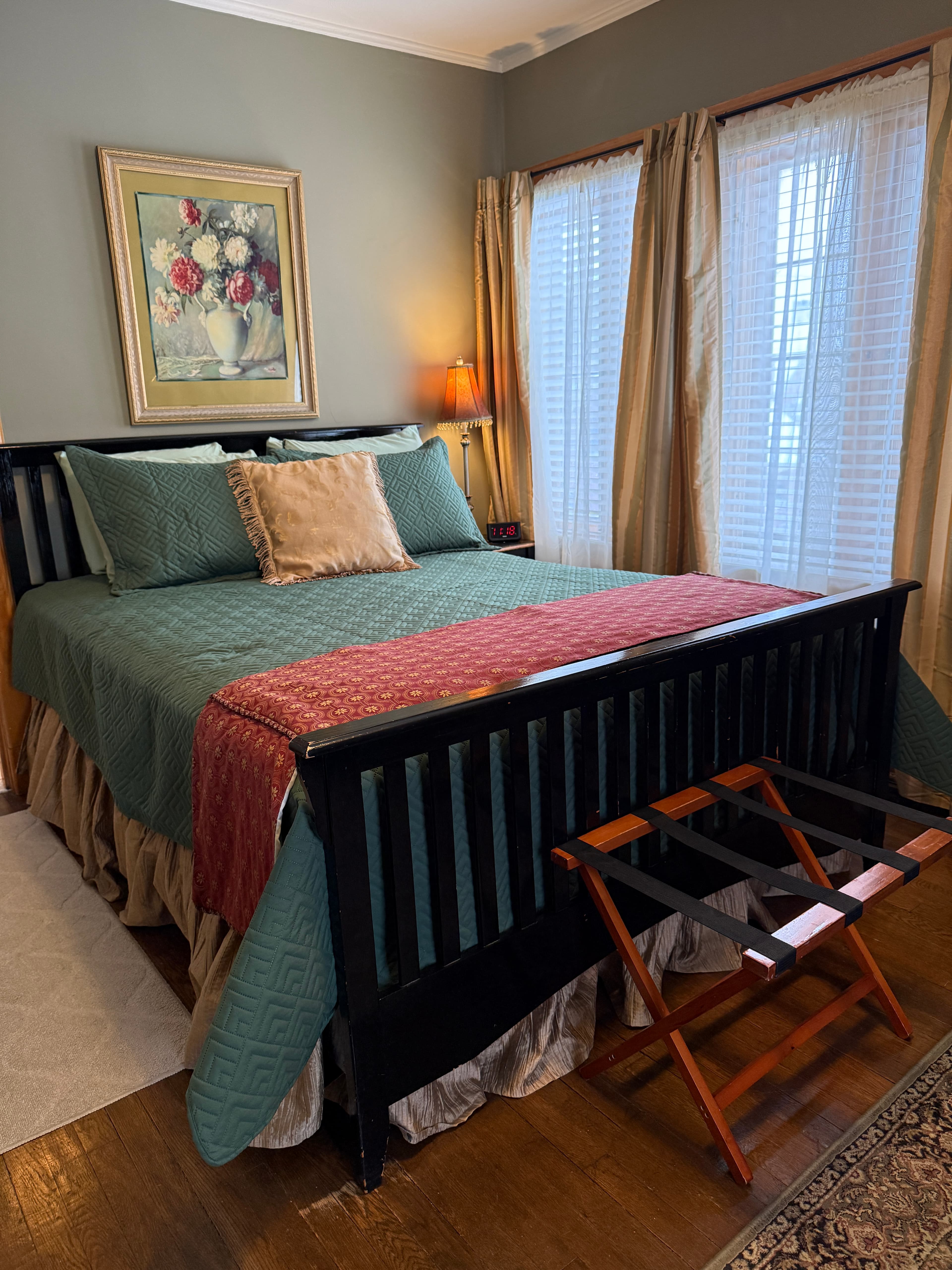 A bedroom with grey-green walls and dark hardwood floors featuring a black slatted bed frame, a teal quilted bedspread with a red patterned throw, and framed floral artwork above the headboard.