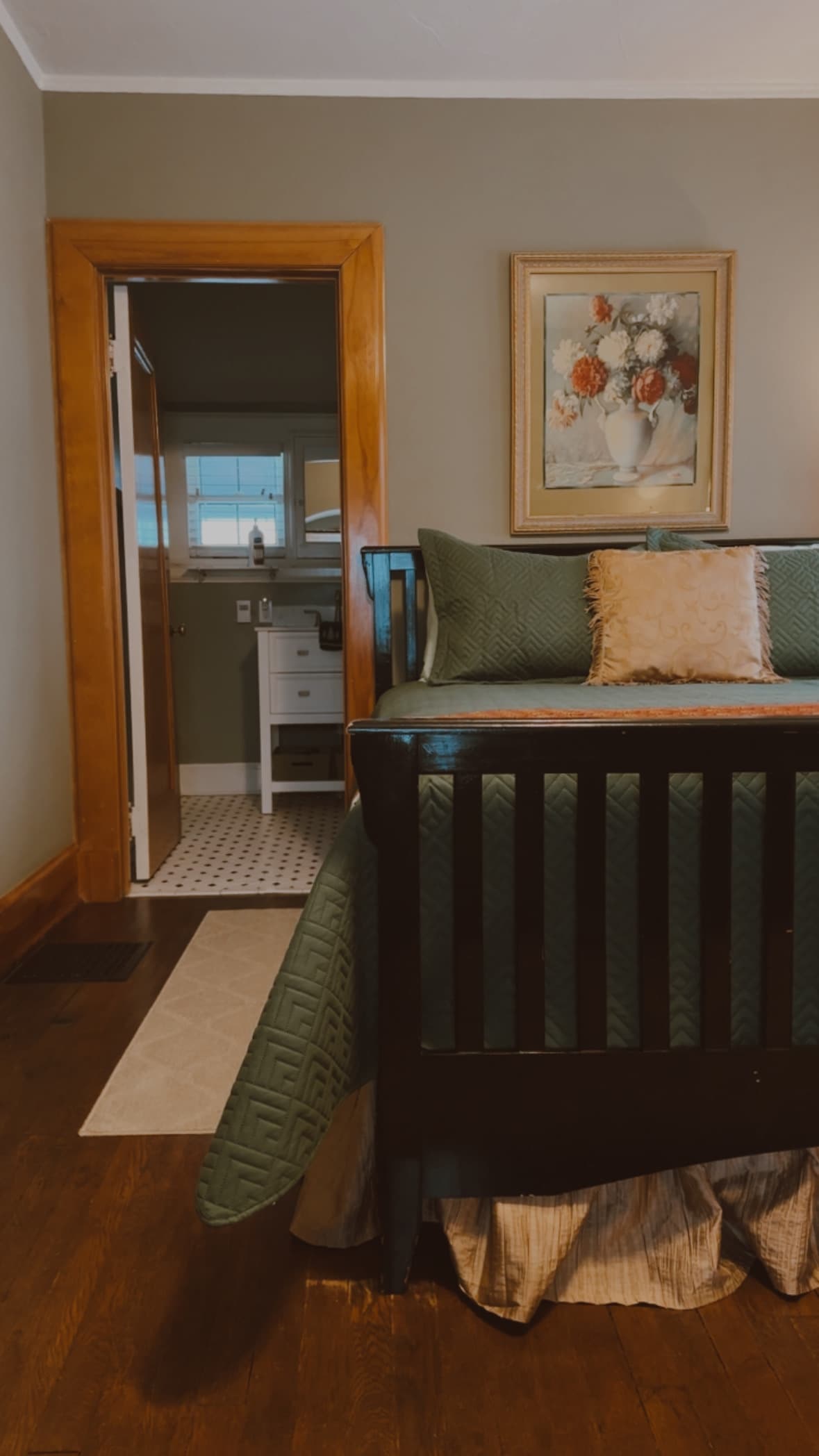 A view from a bedroom with grey-green walls looking through a wooden doorway into a bright bathroom featuring a white vanity with drawers and a white tiled floor.