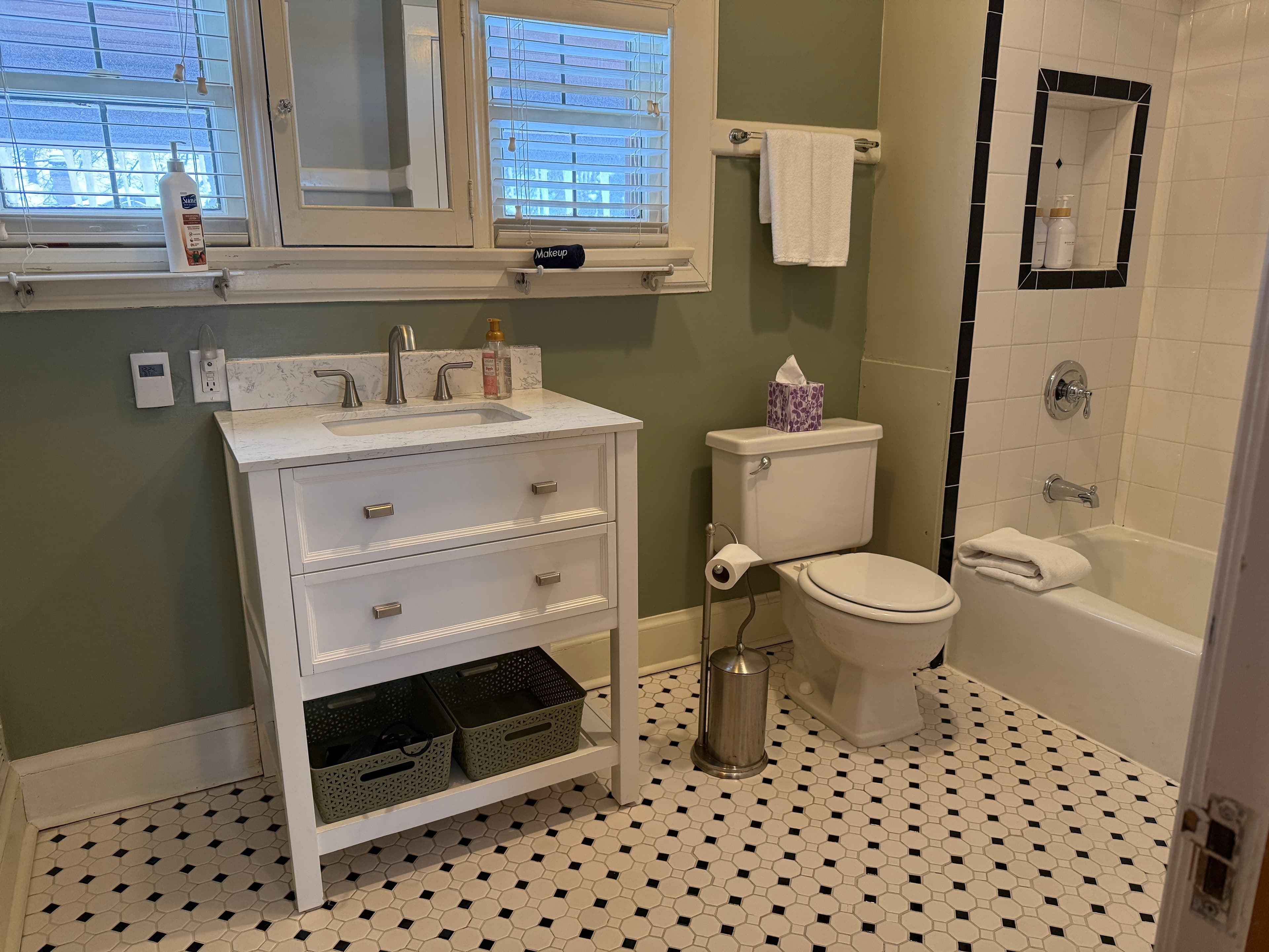 A bright, modern bathroom featuring a white vanity with two drawers and an open shelf, a white toilet, and a bathtub with a white subway-tiled surround. The room is finished with sage green walls and black-and-white patterned tile flooring.
