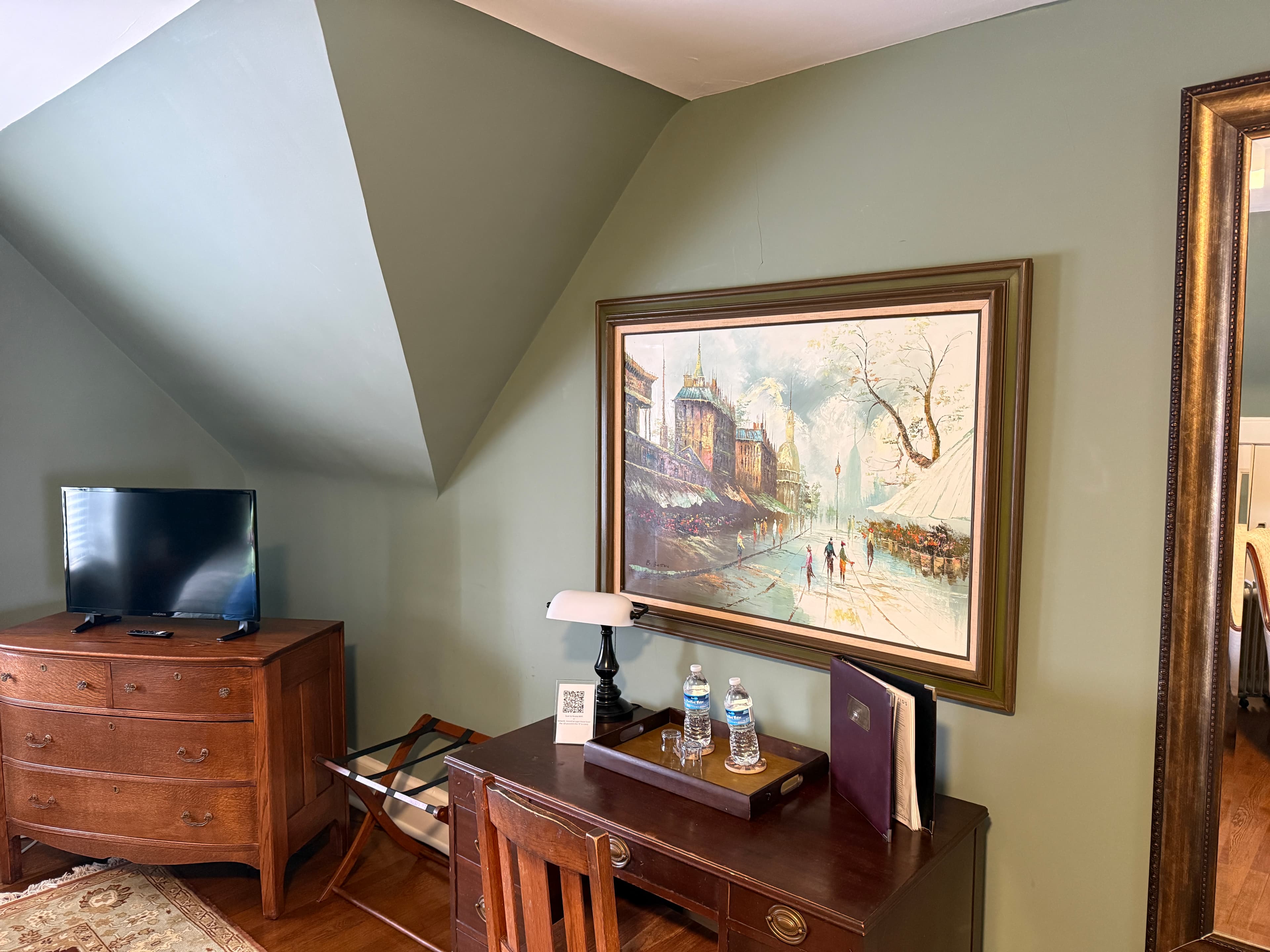 A cozy work area within a room featuring sage green walls and sloped ceilings, showcasing a dark wood desk with a vintage-style lamp, a large gold-framed painting of a European street scene, and a classic wood dresser with a flat-screen TV.