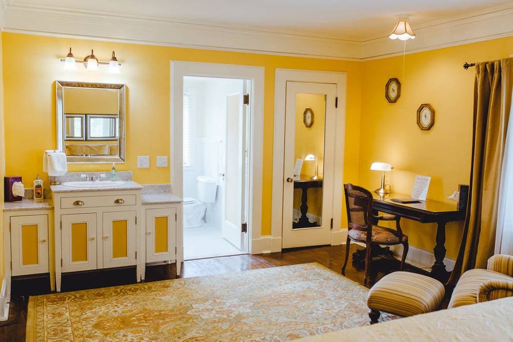 A brightly lit guest room with yellow walls, featuring a white & yellow in-room vanity/sink with a large mirror, a dark wood writing desk with a chair, and a doorway leading into a white-tiled private bathroom.