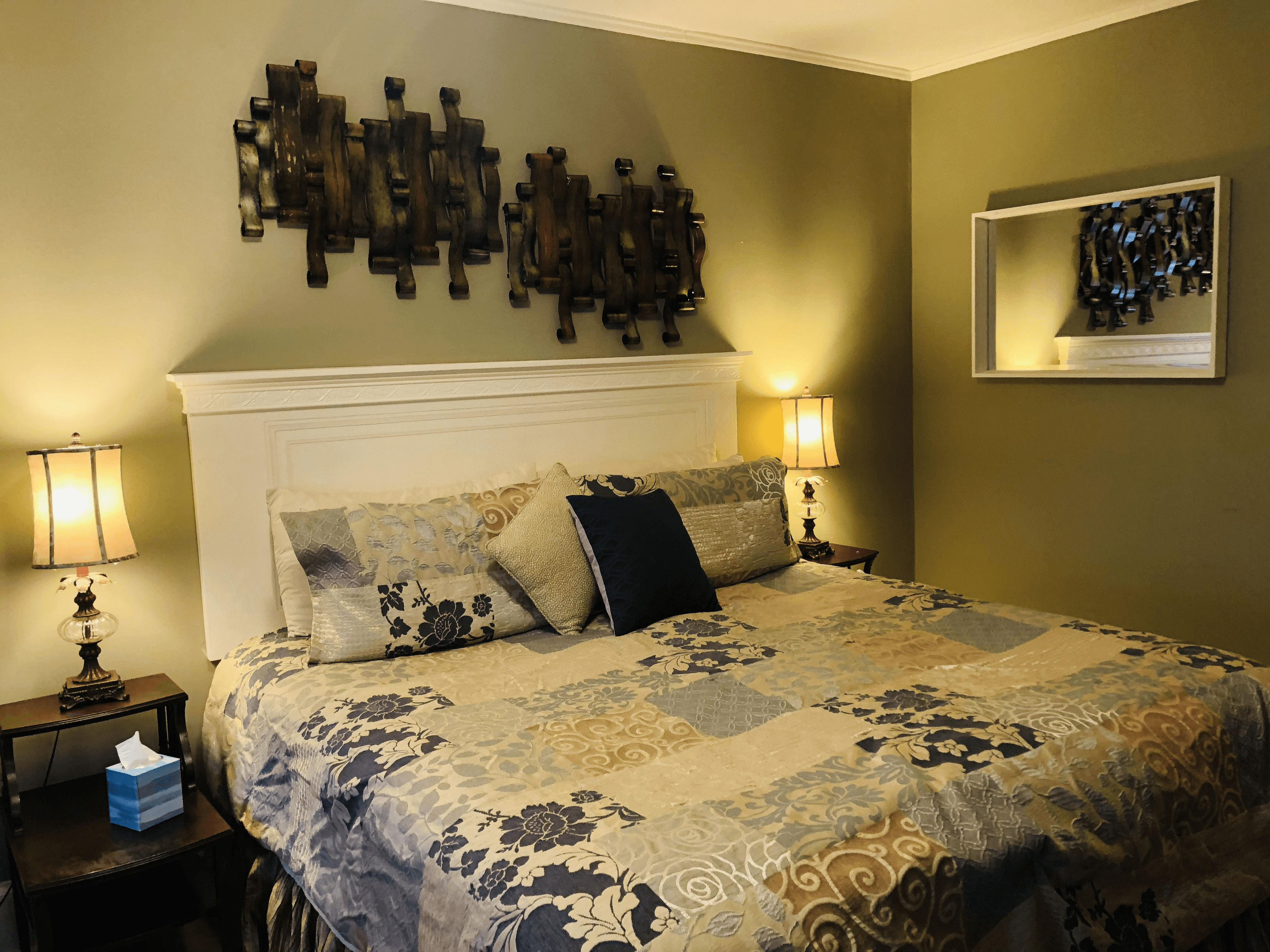 An elegant bedroom with sage green walls featuring a king bed with a white headboard and a blue and beige floral patterned quilt. Intricate dark metal wall art hangs above the bed, flanked by warm lamps on dark wood nightstands.
