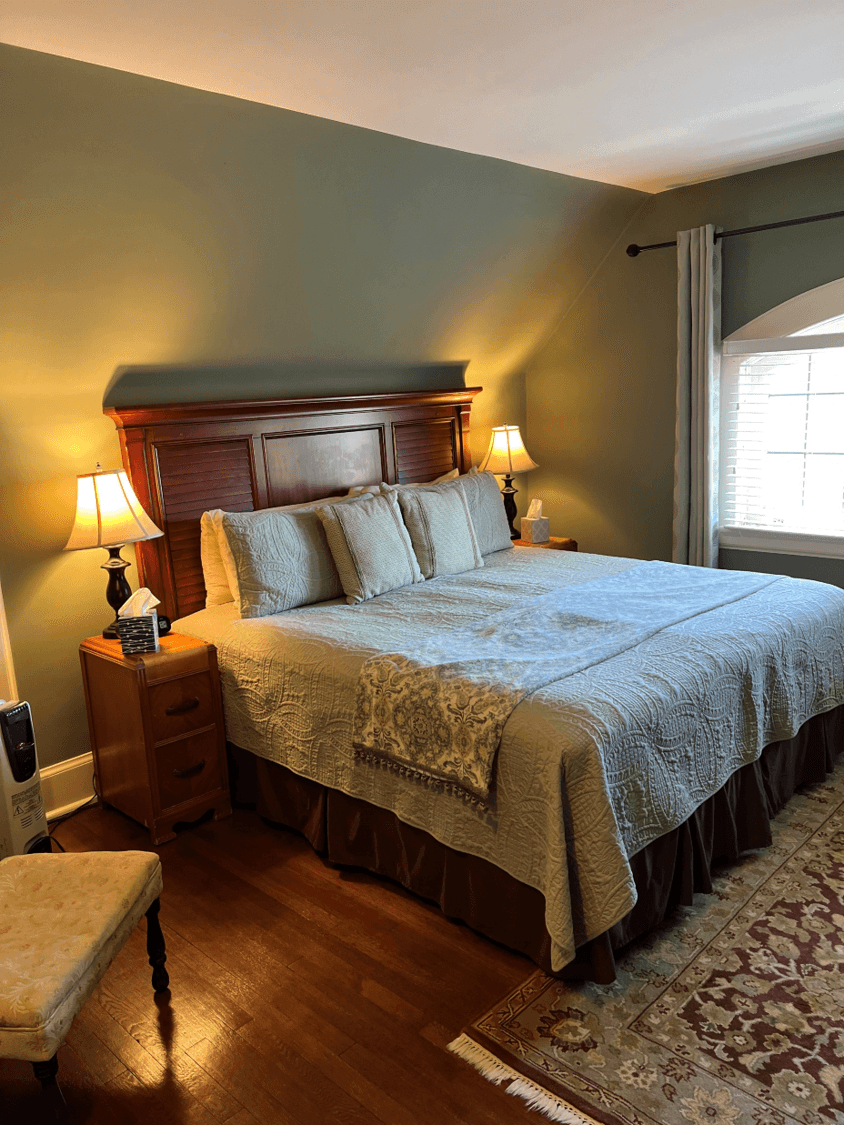 A classic bedroom with sage green walls and dark hardwood floors featuring a large wood-paneled bed with sage green bedding, a damask blanket, and glowing lamps on matching bedside tables.