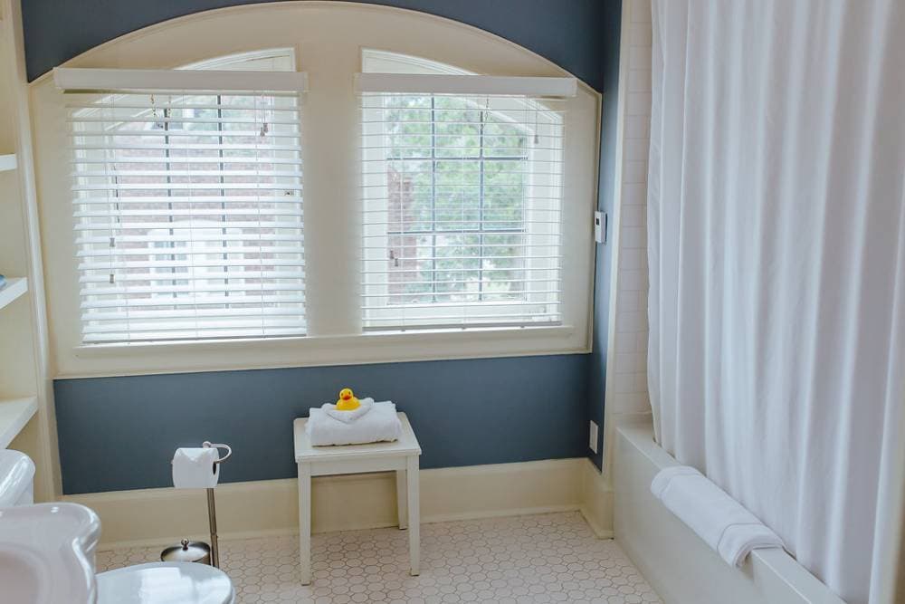A brightly lit bathroom with heated floor, deep blue walls and white trim, featuring two arched windows with white blinds. A white stool sits between the windows, and a bathtub with a white shower curtain is visible on the right.