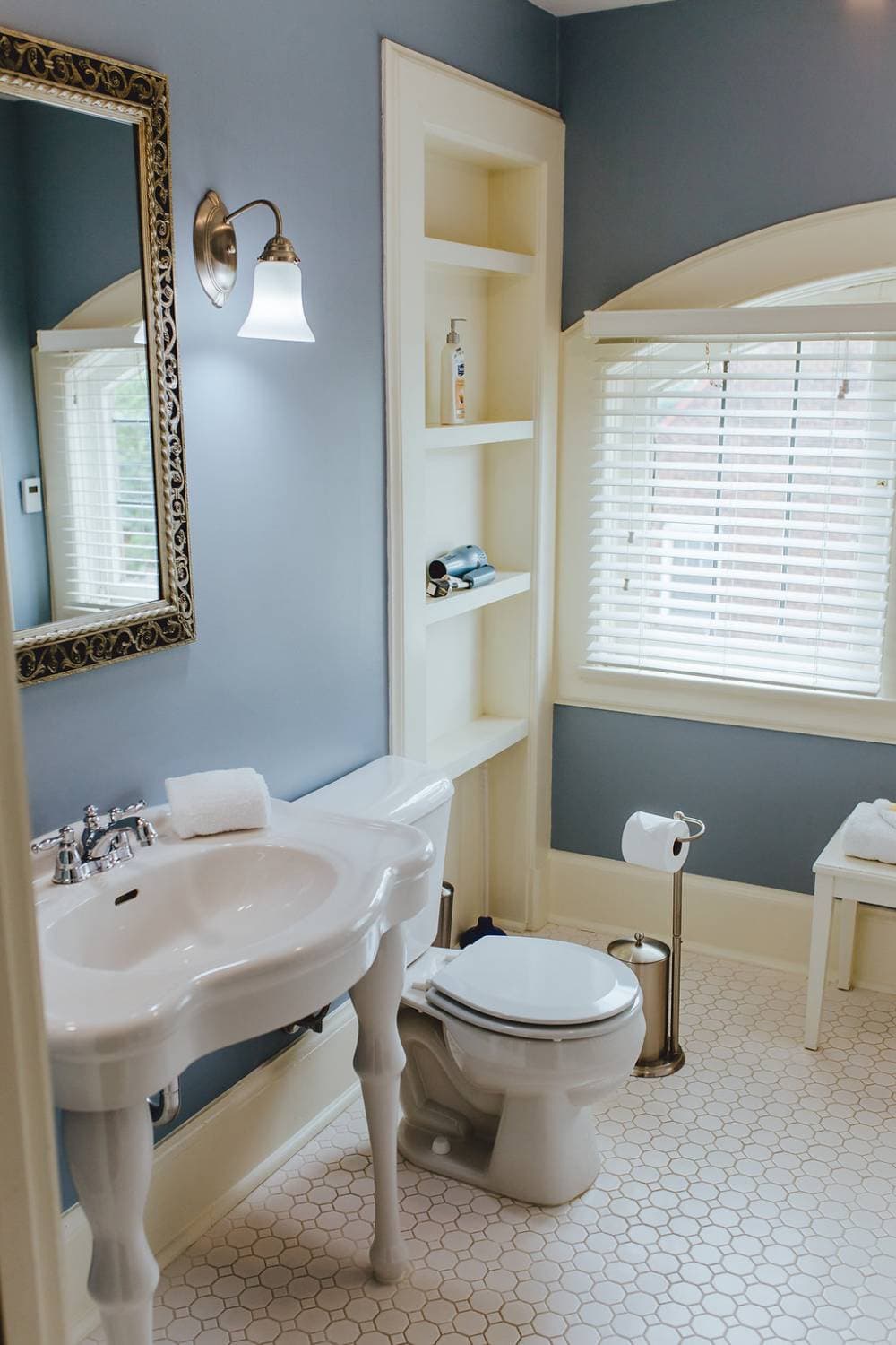 An elegant, vintage-inspired bathroom featuring blue-grey walls, a white porcelain pedestal sink with a framed mirror above, and a white toilet. The space is brightened by an arched window with white blinds and features white penny tile heated floor.