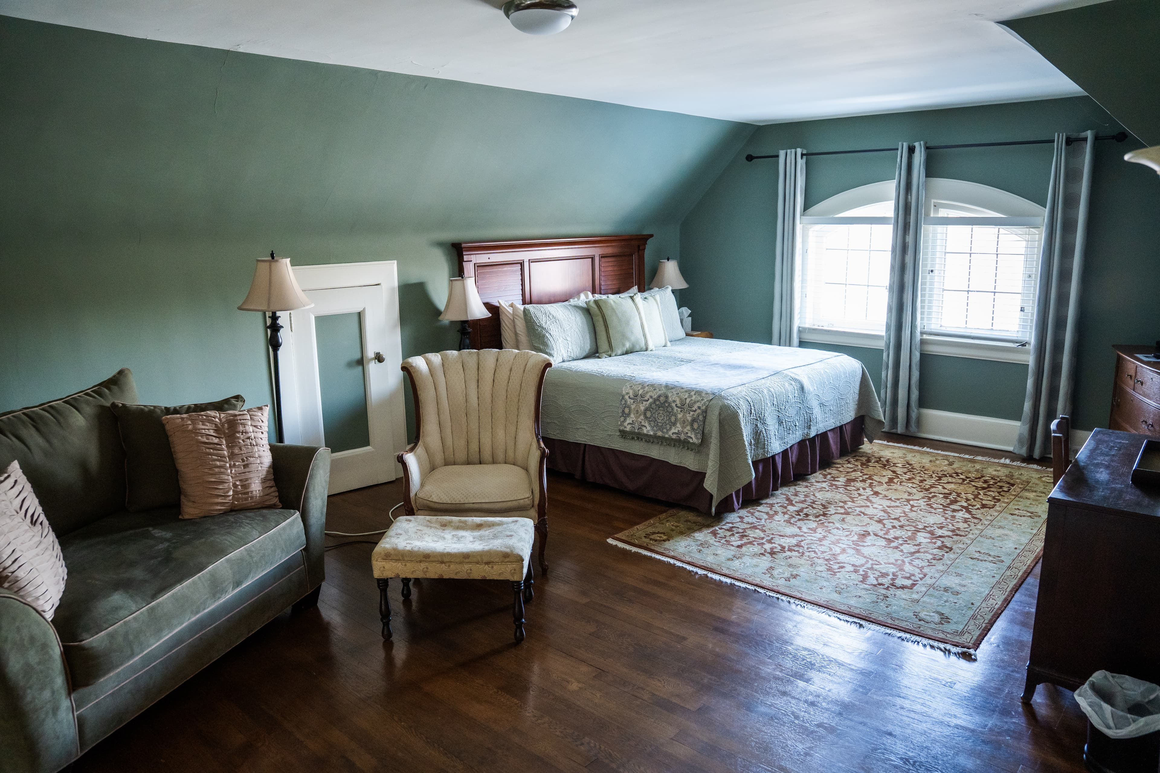 A spacious bedroom with sage green walls and a sloped ceiling, featuring a large wood-framed bed with sage bedding, a comfortable green sofa, and a cream wingback chair with a matching ottoman.