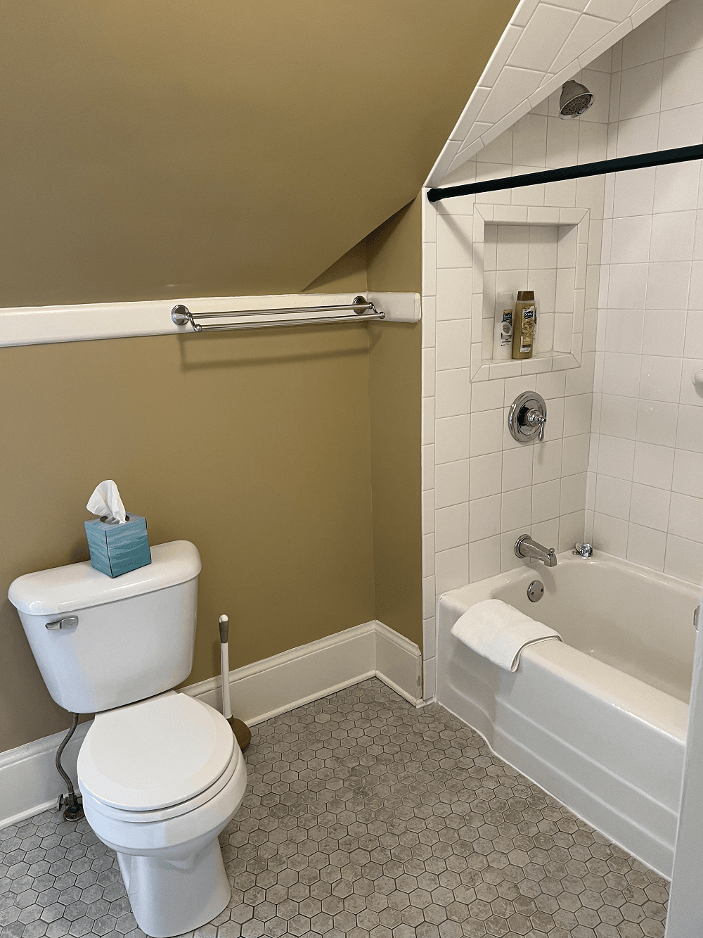 A private bathroom featuring tan walls and grey penny tile heated floor. It includes a white toilet and a built-in white bathtub and shower combination under a sloped ceiling, with white wall tiles and a black shower rod.
