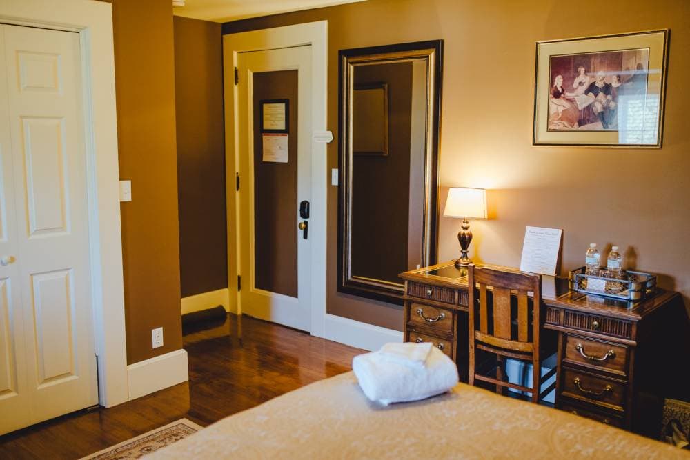 A classic bedroom featuring a small dark wood writing desk with a chair, a large wall mirror, and a framed picture on tan walls. A bed with beige quilted linens is visible in the foreground.