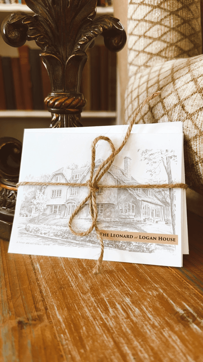 A decorative card featuring a sketch of the Leonard at Logan House, tied with twine, placed on a wooden table.