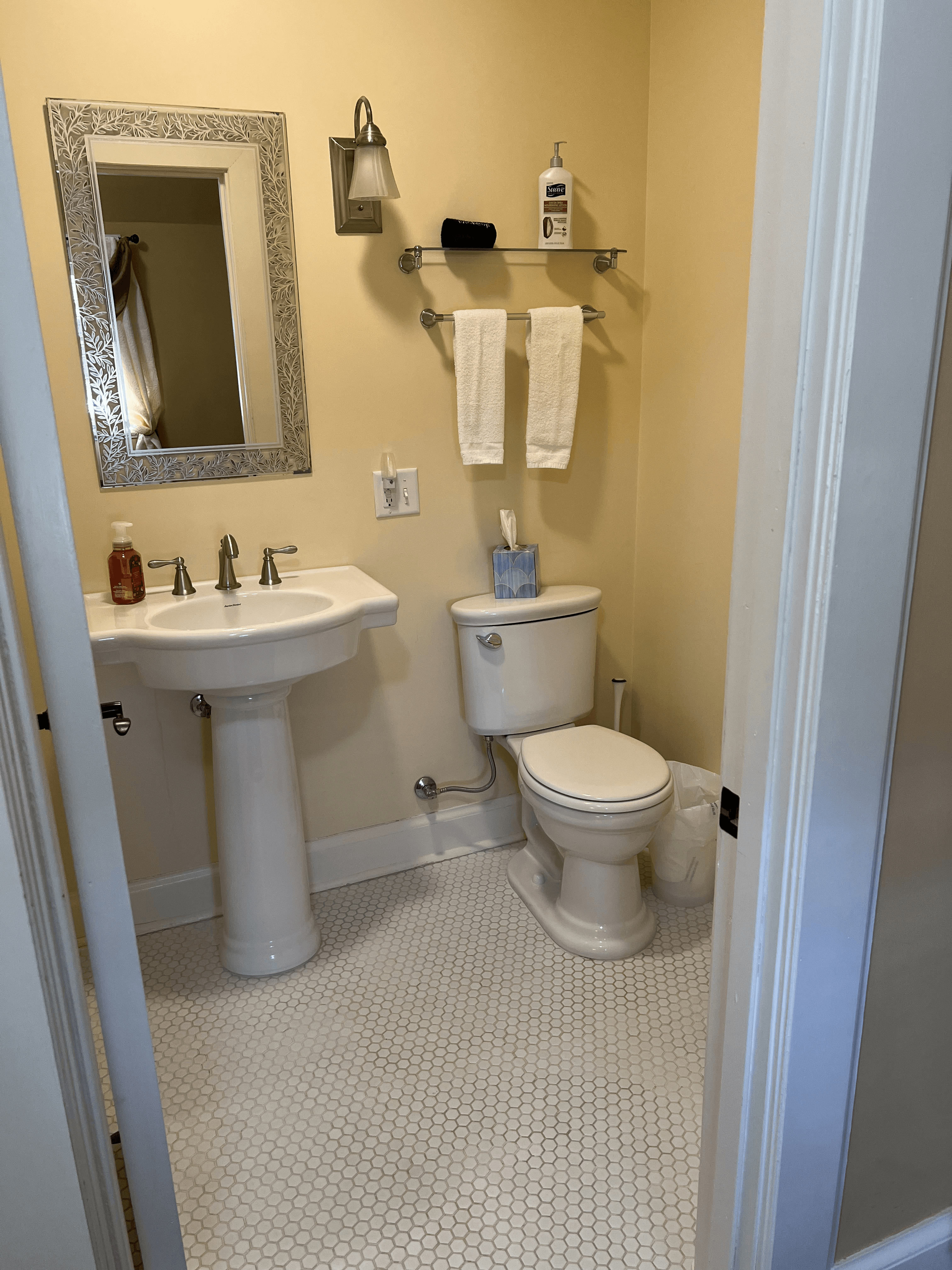 A brightly lit bathroom with yellow walls and white penny tile flooring, featuring a classic white pedestal sink, a silver-framed mirror, a toilet, and fresh white towels on a glass wall shelf.