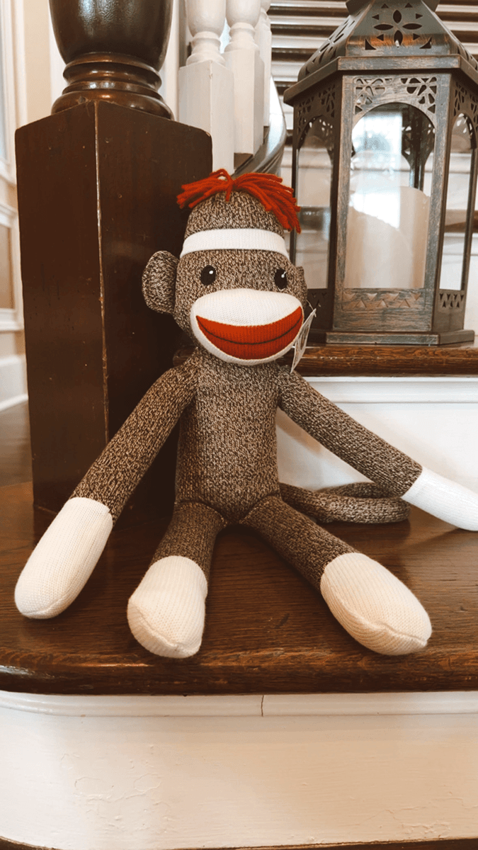 A cheerful sock monkey toy with a red mouth and hair sits on a wooden surface next to a lantern.