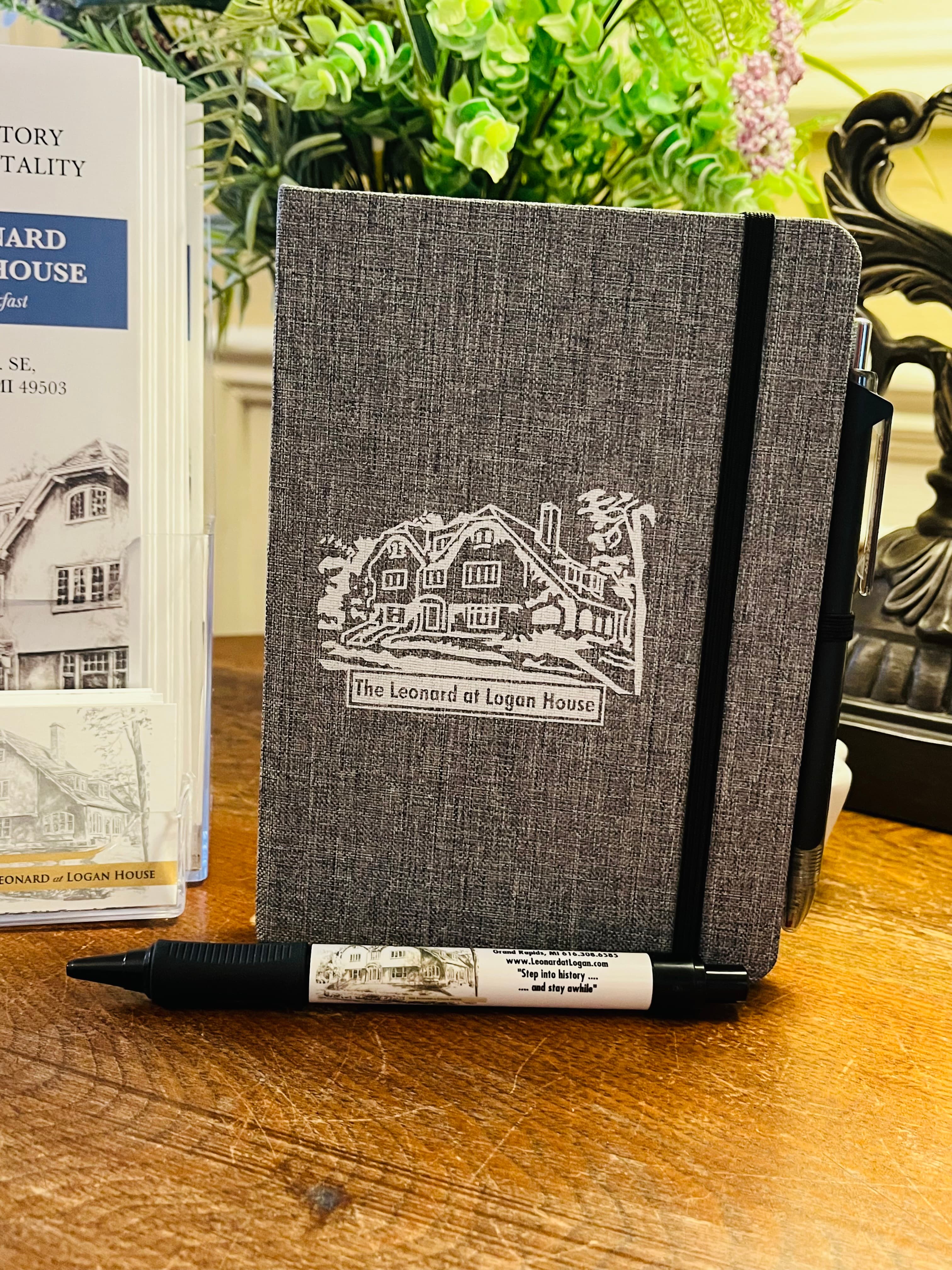 A gray notebook with "The Leonard at Logan House" printed on it, accompanied by a pen, sits on a wooden table near a brochure display.
