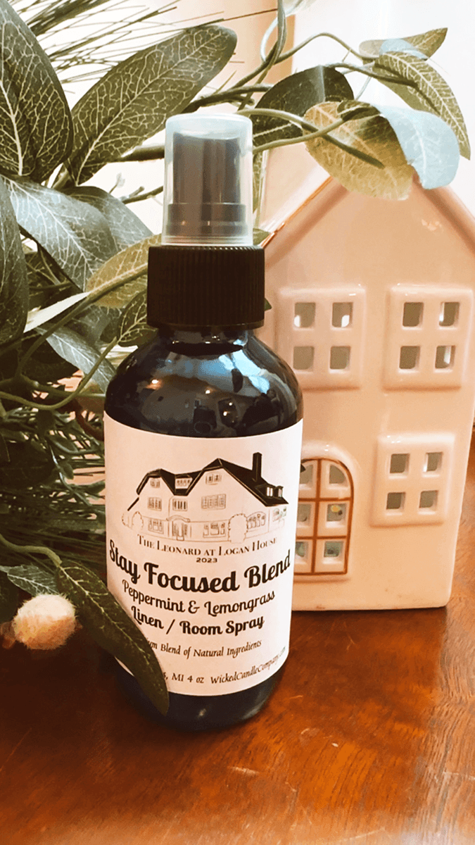 A bottle of "Stay Focused Blend" peppermint and lemongrass room spray is displayed next to a decorative ceramic house and green foliage.