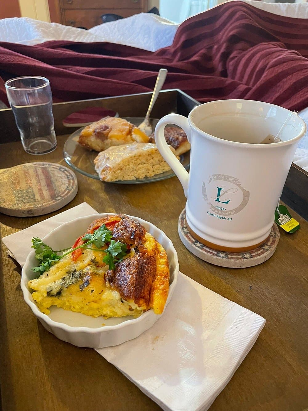 A cozy breakfast tray featuring an omelette, scones, a glass of water, and a mug of tea next to a tucked-in bed.