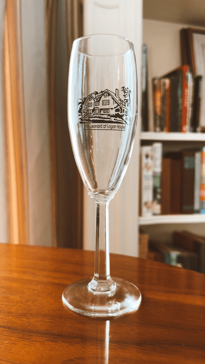 A clear champagne flute with a logo of "The Leonard at Logan House" sits on a wooden table.