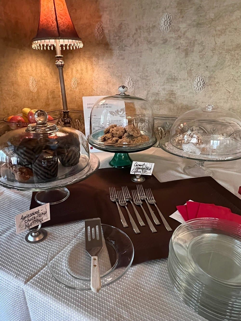 A display of assorted baked goods under glass cloches, next to plates and utensils, illuminated by a vintage lamp.