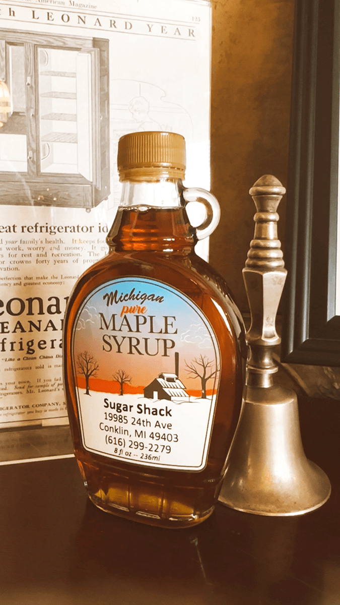 A bottle of Michigan pure maple syrup labeled "Sugar Shack" sits beside a silver bell against a vintage backdrop.