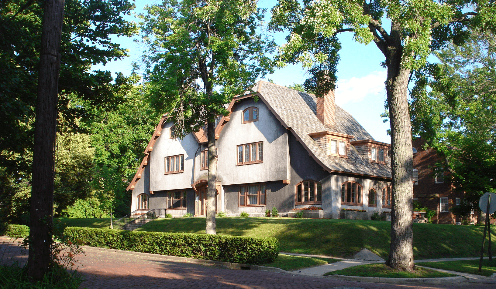 A large, unique house surrounded by trees and green lawn on a residential street.