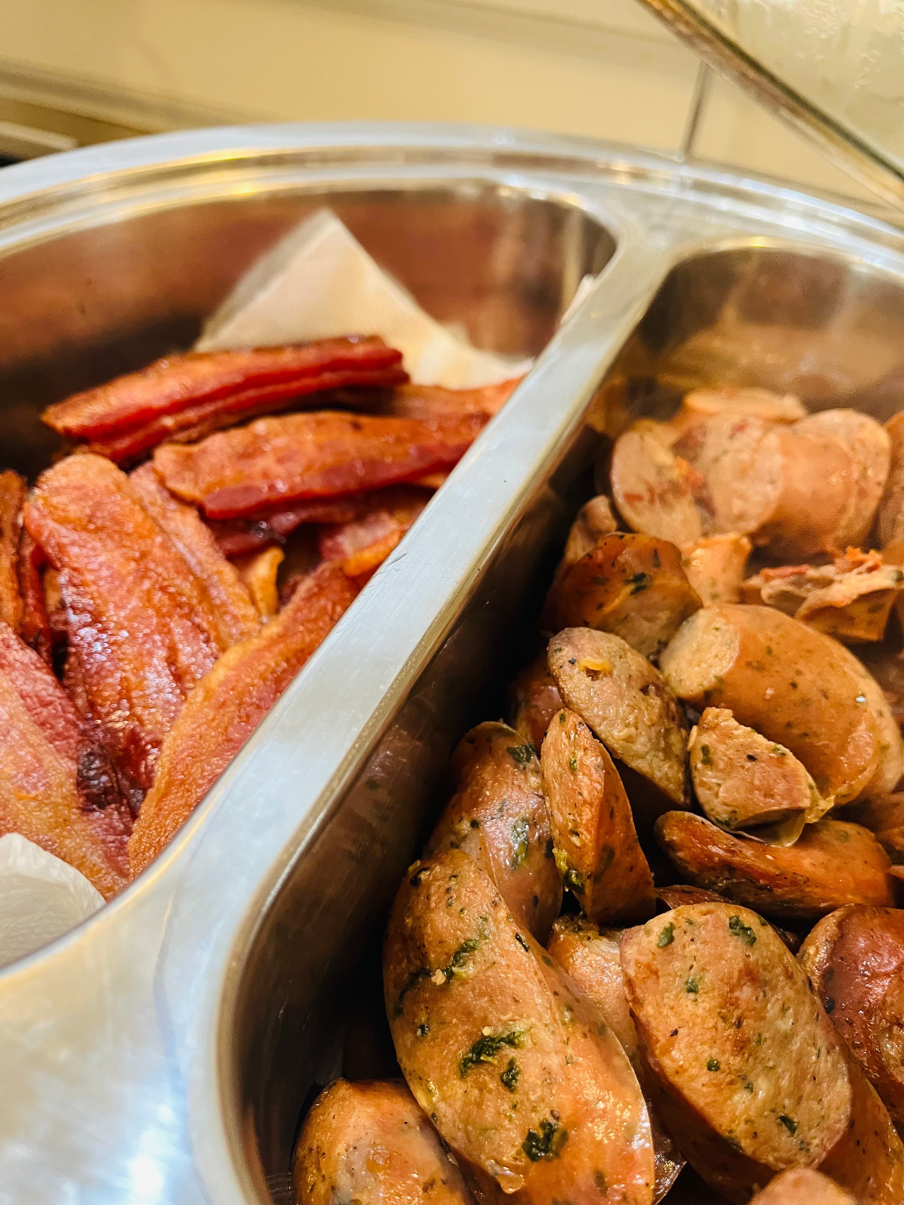A close-up of a serving tray divided into two sections, one filled with crispy bacon strips and the other with sliced sausages.