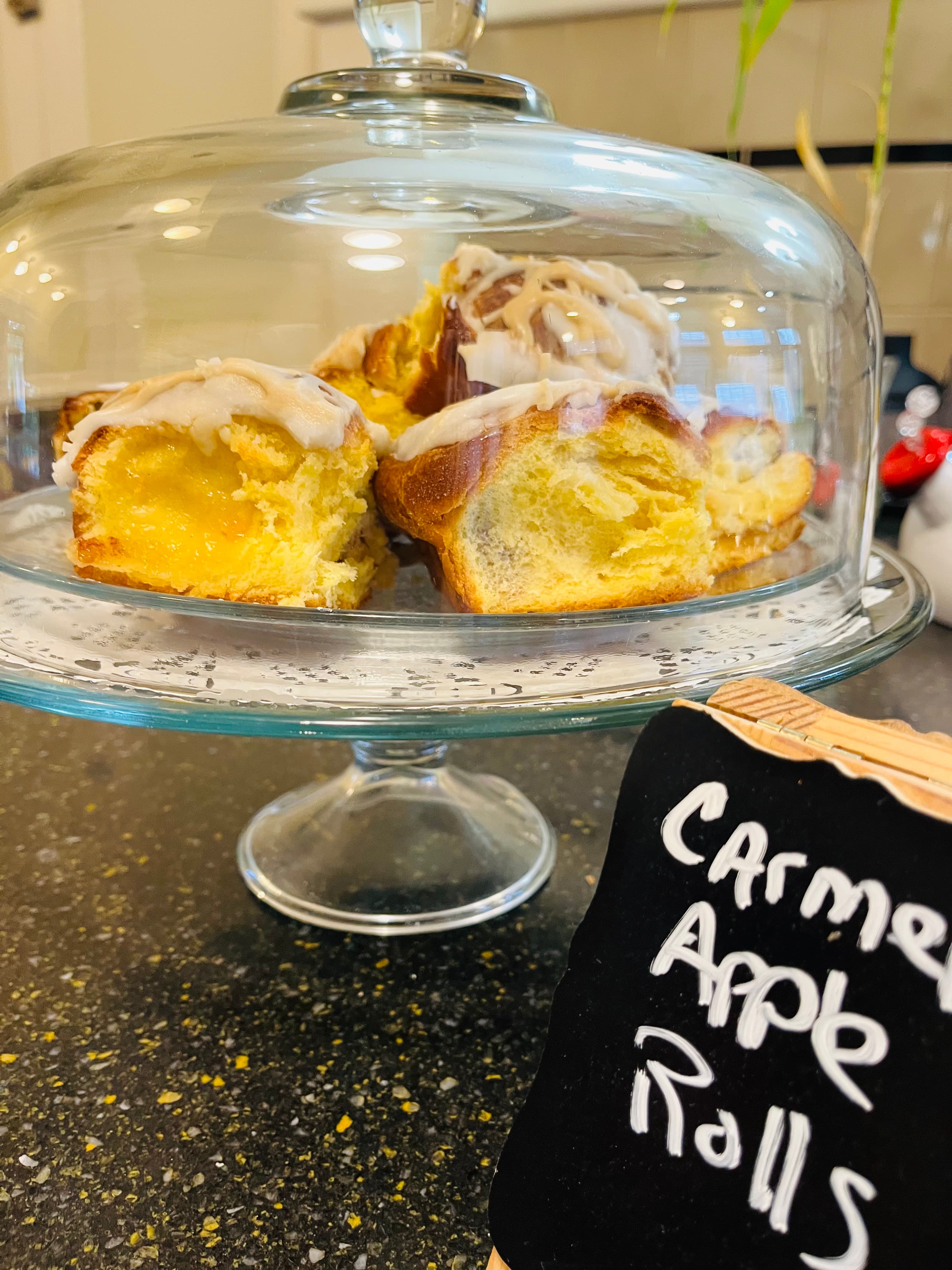 A glass dome displays sliced caramel apple rolls on a cake stand, with a sign labeled "Caramel Apple Rolls."