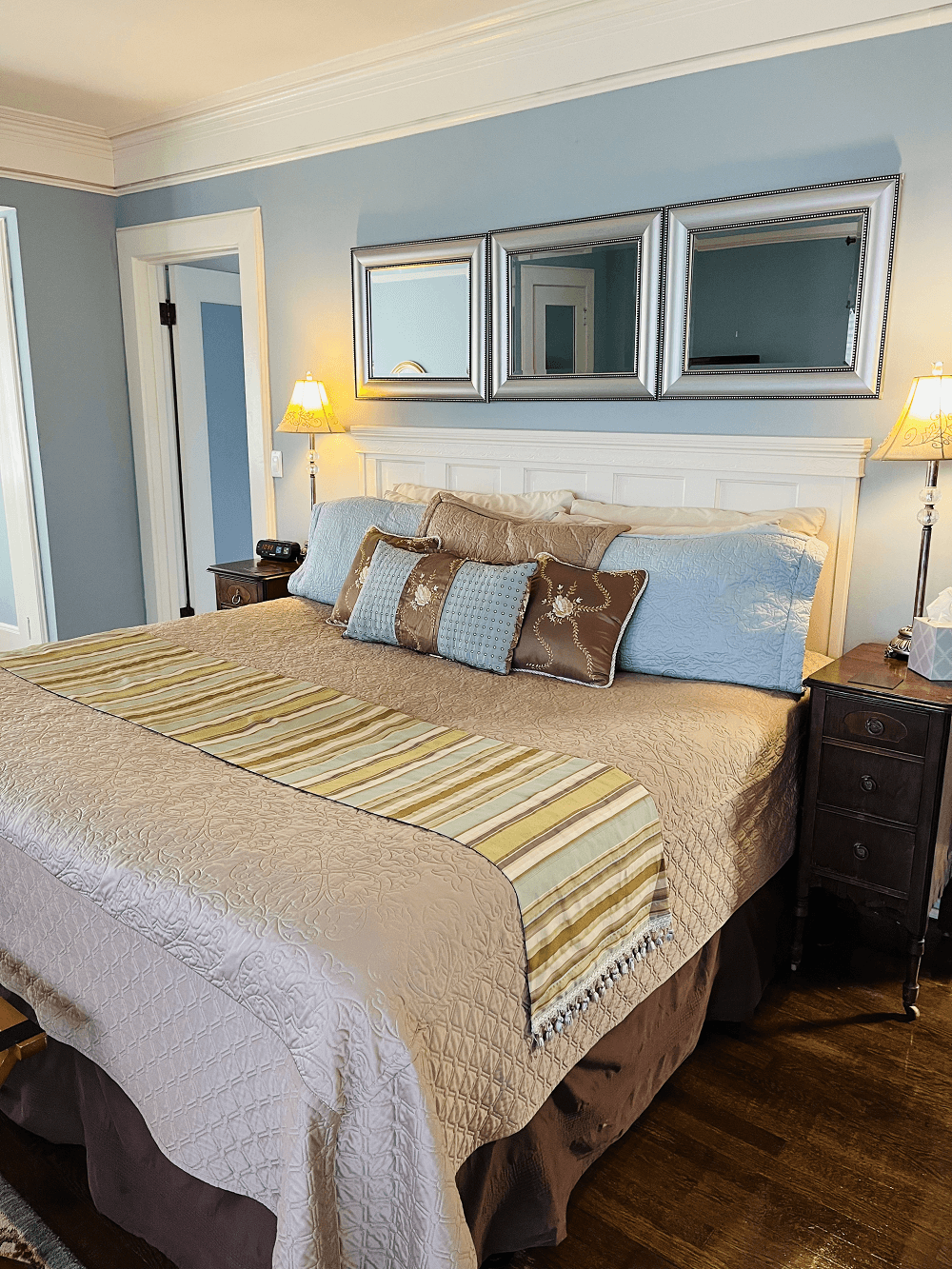 A serene bedroom with light blue walls featuring a king bed with a white headboard, a beige quilted bedspread, and a striped runner. Three square mirrors hang above the bed, flanked by classic bedside lamps.