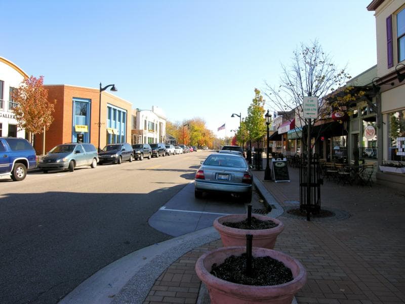 A quiet downtown street lined with trees, parked cars, and shops under a clear blue sky.