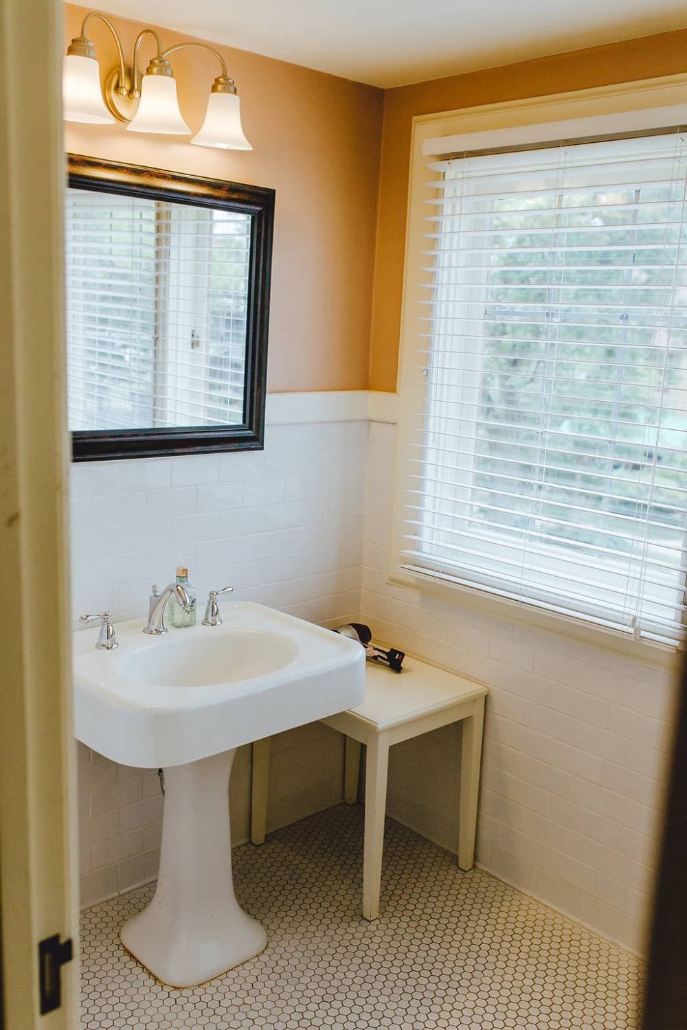 A brightly lit bathroom featuring a classic white pedestal sink, a small white side table, and a large window with white blinds. A dark-framed mirror and a three-bulb light fixture hang on the tan wall.