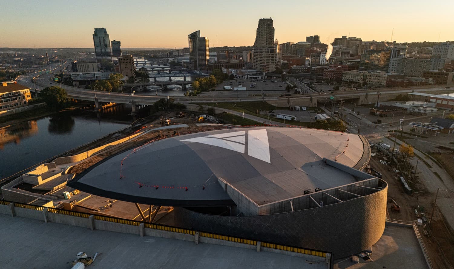 Aerial view of a modern building with a distinctive triangular roof, surrounded by a city skyline at sunset.