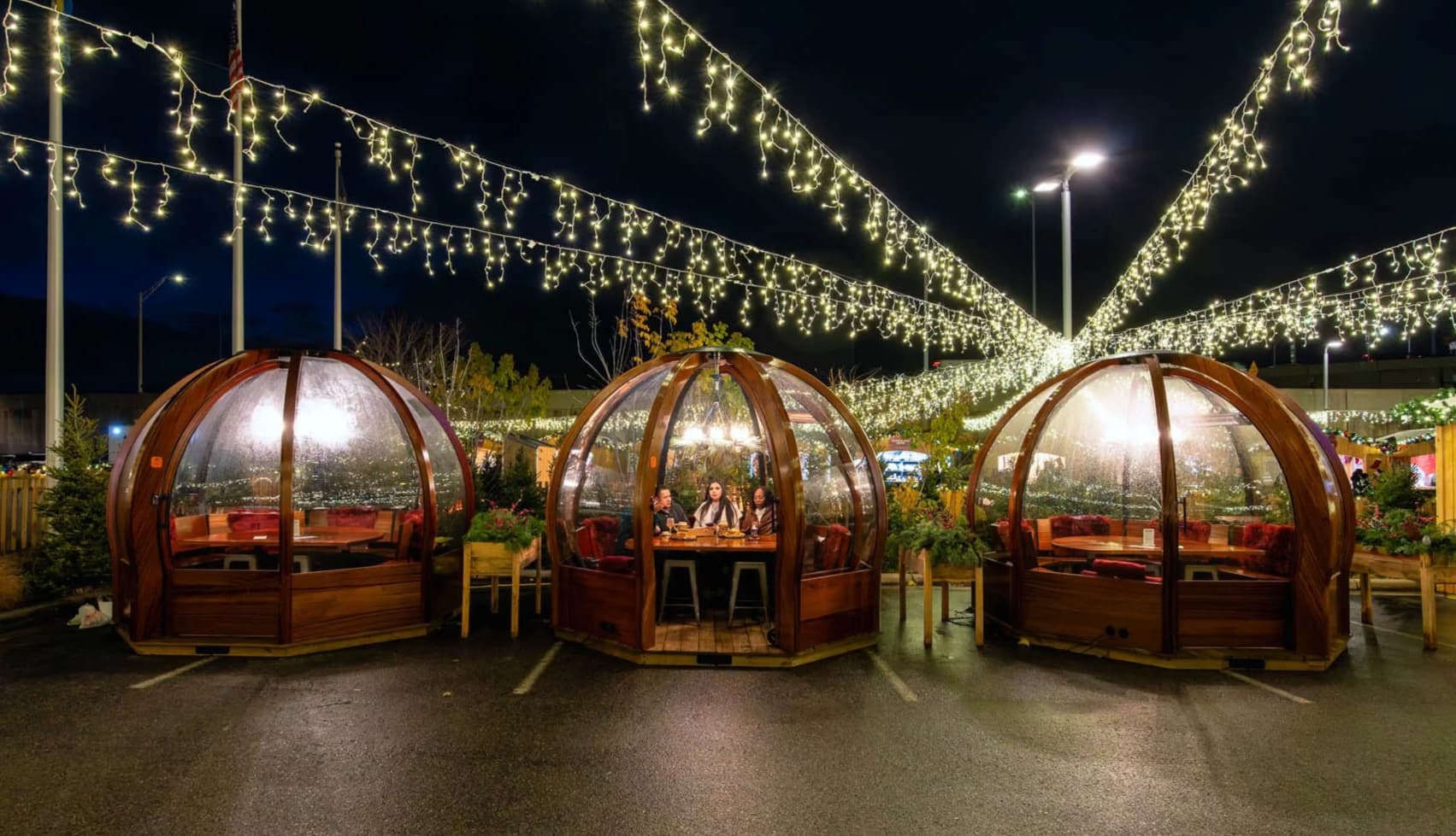 Dining igloos lit with string lights at night. Two wooden igloos house people dining, surrounded by greenery, creating a cozy, festive atmosphere.