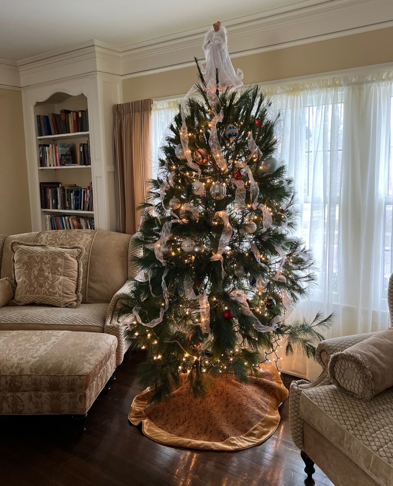 A decorated Christmas tree adorned with lights, white ribbons, and ornaments stands in a cozy living room. Soft light filters through the window, casting a warm, inviting glow.