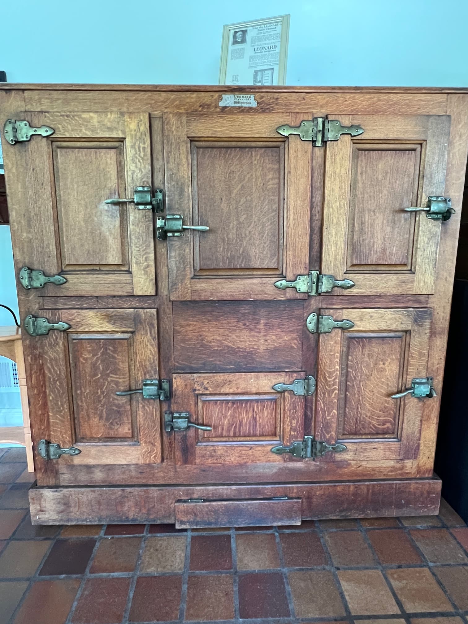 Antique wooden icebox with five paneled doors, brass hinges, and latches, set against a light blue wall on a brown tiled floor. Archival paper is displayed on top.