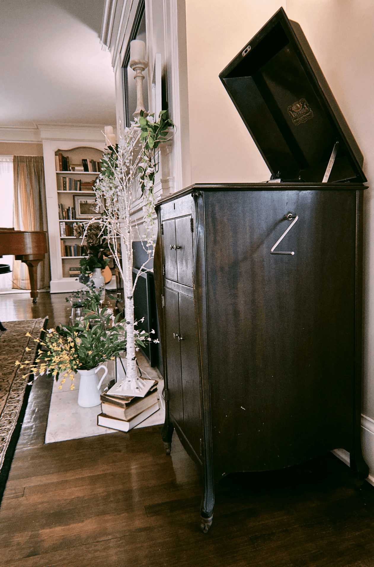 Vintage wooden cabinet with an open lid beside a white decorative tree and flowers. Background shows a cozy room with bookshelves and a grand piano.